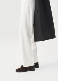 Scarpe con tacco in suede marrone con punta arrotondata, abbinate a pantaloni bianchi larghi e un cappotto nero, che mostrano un sottile contrasto di texture.