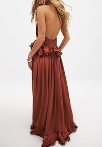 Robe en satin couleur rouille avec un dos nu, une taille smockée et des accents volantés au niveau du buste et de l'ourlet. Design fluide, longueur sol.