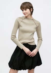 Πλεκτό μπεζ πουλόβερ με ριγέ υφή και λεπτομέρειες με κουμπιά στο πλάι, συνδυασμένο με μαύρη φούστα με καμπάνα. Το σύνολο έχει εφαρμοστή γραμμή και υφές με υφή.