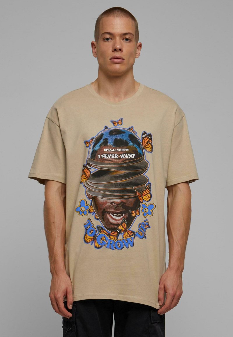 Camiseta de algodón beige con estampado gráfico de un rostro con sombreros, rodeado de flores azules y patrones de mariposas naranjas. Diseño de corte holgado.