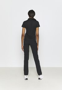 Black short-sleeve polo and fitted black pants. Smooth fabric with minimalist design. Model wearing white athletic shoes, viewed from the back.