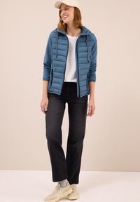 Gevoerde blauwe jas met een capuchon, ritsluiting, gecombineerd met een wit t-shirt en zwarte broek. Tan sneakers met gestructureerde zolen. Neutrale achtergrond.