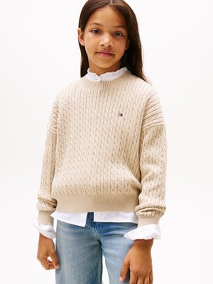 Beige cable-knit pulóver, bordás mandzsettával és szegéllyel, fehér galléros ing felett viselve. Lazán szabott. Kis logó akcentus a mellkason.