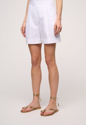 Shorts blancs ajustés avec un devant plat et des jambes larges. Associés à des sandales dorées à lanières avec un détail en nœud et des liens fins autour de la cheville.