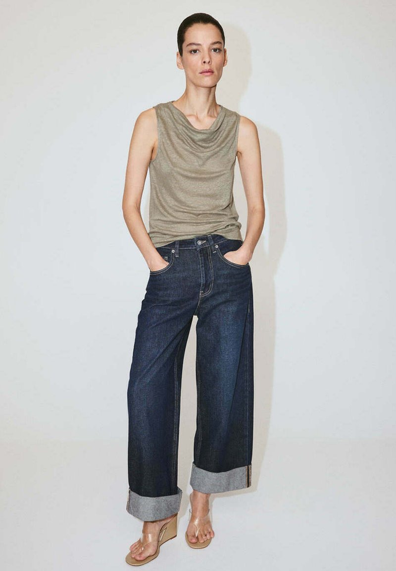 Femme les mains dans les poches, portant un haut beige sans manches, un pantalon large en jean foncé avec revers, et des sandales compensées à brides transparentes, debout devant un fond uni.