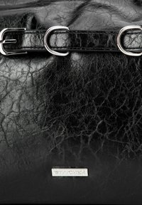 Czarna skórzana torebka o pomarszczonej fakturze, z metalowymi elementami w kolorze srebrnym oraz wyraźną metalową plakietką z logo. Uzupełniona zapięciem w stylu paska.