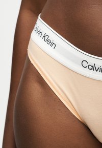Κοντινό πλάνο μέσης με σκούρο χρώμα δέρματος που φοράει μπεζ εσώρουχα Calvin Klein με λευκή μέση που αναγράφει το λογότυπο της μάρκας με μαύρα γράμματα.