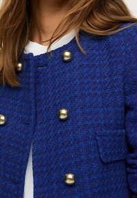 Manteau bleu texturé avec un motif tissé, doté de grands boutons dorés et d'une petite poche à l'avant. Tissu doux, design structuré.