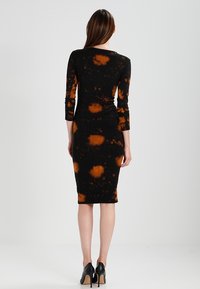 Černé šaty s dlouhými rukávy a oranžovými skvrnami tie-dye, přiléhavý střih, do poloviny stehen, vyrobené z elastického materiálu, v kombinaci s černými lodičkami na vysokém podpatku.