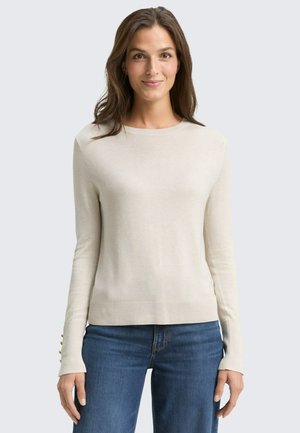 Helles beiges Langarmpullover mit geripptem Saum und Knopfverzierungen an den Ärmeln. Kombiniert mit blauen Jeans aus glattem Material.