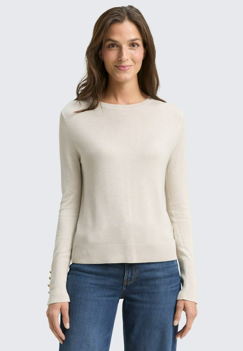 Lichtbeige lange mouwen sweater met ribbeltrim, voorzien van knoopaccenten op de mouwen. Gecombineerd met een blauwe spijkerbroek, gladde textuur.