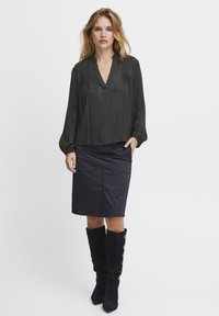 Blusa nera con texture, scollatura a V e dettagli plissettati, abbinata a una gonna a matita scura con tasche frontali e stivali neri al ginocchio.