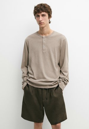 Jonge man die een beige langarm henley-shirt en donkerolijfgroene shorts draagt, staand met handen in zijn zakken tegen een witte achtergrond.