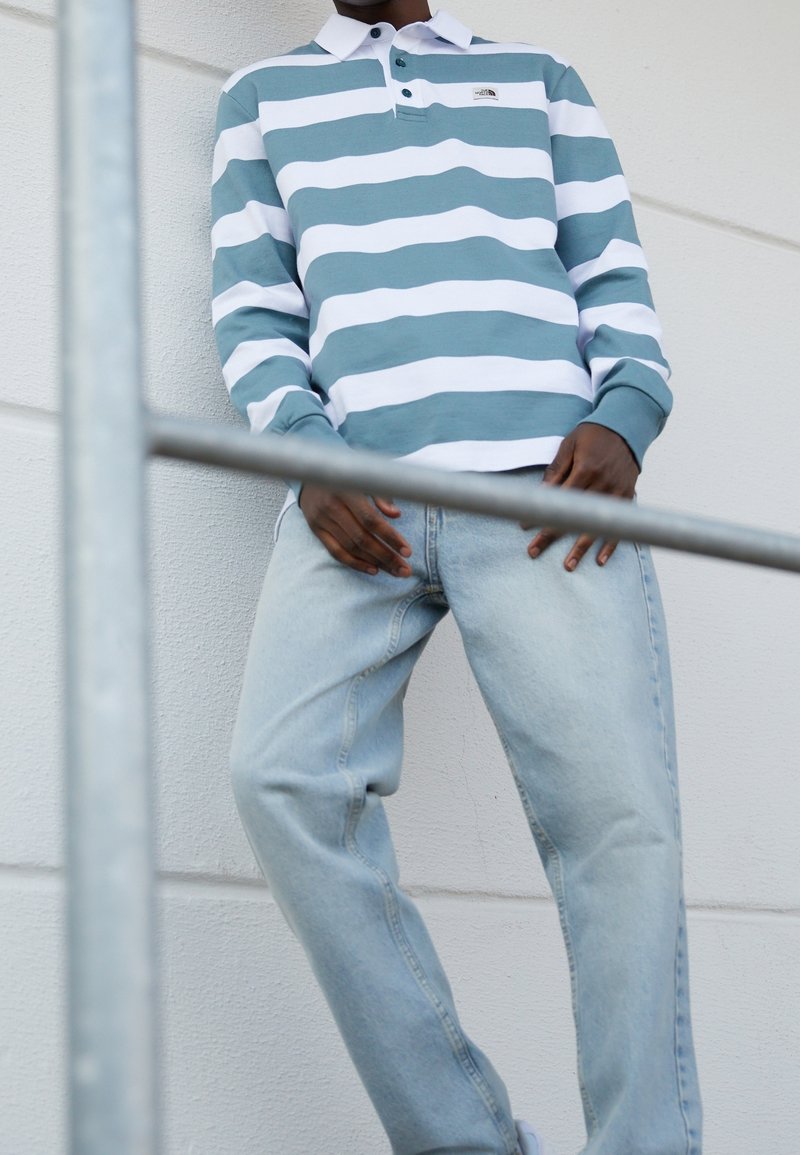 Personne portant une chemise à manches longues à rayures horizontales bleu et blanc et un jean bleu clair, debout contre un mur blanc avec des barres métalliques devant.