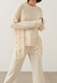 Neulepaita kermanvärisenä, ribbauslahkeilla ja sivuilla fringed-yksityiskohdilla, yhdistettynä vaaleanbeigeihin leveälahkeisiin housuihin.
