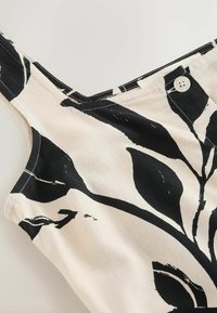 Haut beige sans manches avec de larges bretelles, motifs abstraits de feuilles noires et un bouton blanc près de l'encolure sur fond clair.