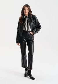 Czarna skórzana kurtka typu cropped, w paski koszula z guzikiem, dopasowane czarne skórzane spodnie oraz botki z błyszczącym wykończeniem. Prosty, elegancki design.
