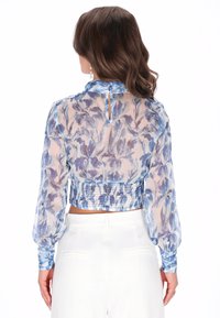 Bloes met bloemenprint van doorzichtig blauwe stof, lange pofmouwen en een gesmokte zoom. Heeft een hoge kraag en een knoopsluiting op de rug.