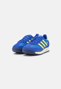 Chaussures de sport bleues avec des rayures jaunes, en tissu et en daim, présentant une tige texturée, des lacets et une semelle en caoutchouc à profil bas.