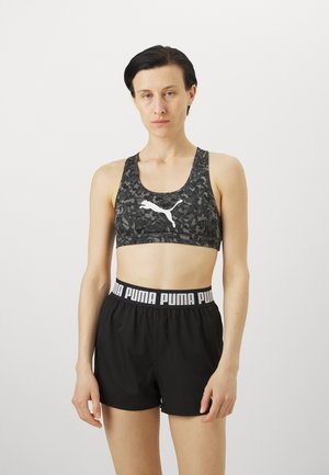 Svart kamuflasje sports-BH med hvit logo, kombinert med svarte shorts med en hvit merkebånd i livet. Glatt tekstur, atletisk design.