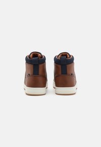 Brązowe skórzane sneakersy typu high-top z granatowymi wyściółkami i białymi gumowymi podeszwami widoczne z tyłu na białym tle.