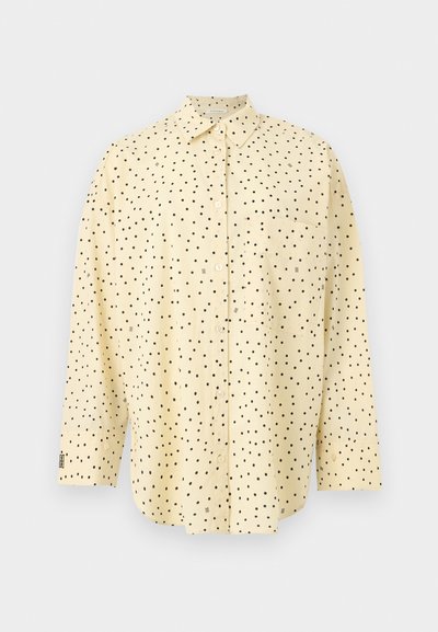 Chemise à manches longues de couleur jaune clair avec des boutons et des pois noirs, dotée d'une poche poitrine unique, présentée sur un fond uni.