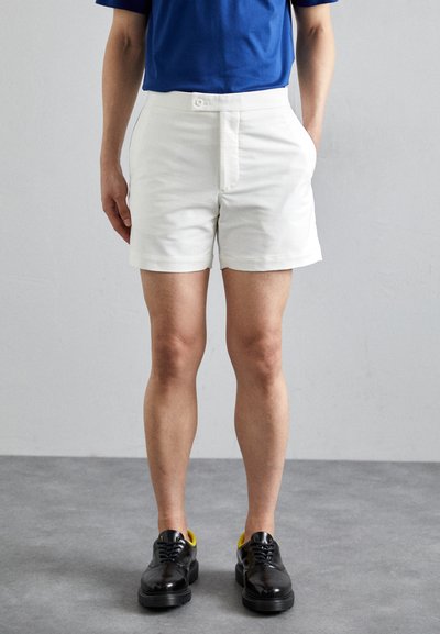 Shorts din bumbac alb, cu croială dreaptă, buzunare frontale și închidere cu buton, asociați cu pantofi negri cu accente galbene.
