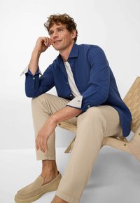 Blue linen shirt with contrasting light cuffs, cream t-shirt underneath, beige chinos, and beige slip-on shoes, seated on a wooden chair.