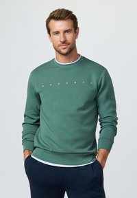 Man draagt een groene Hackett sweater over een wit shirt, met marineblauwe broek, staand met handen in de zakken tegen een effen achtergrond.