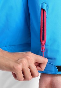 Blaue Jacke mit strukturiertem Material, ausgestattet mit einem pinken Reißverschluss an den Ärmeln und einem schwarzen Klettverschluss am Bündchen. Eine Hand passt das Bündchen an.