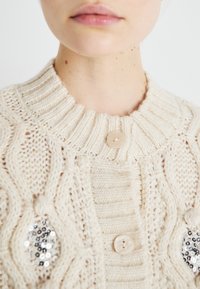 Kremfarget strikket cardigan med ribbestrikket krage, med en stor knapp og teksturerte mønstre, akkompagnert av paljett-detaljer.
