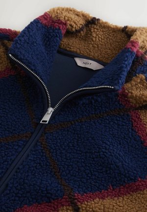 Fleece-tyylinen vetoketjulla varustettu takki, jossa on sininen ja ruskea geometrinen kuvio, kontrastina punaiset yksityiskohdat ja metallic-värinen vetoketju kauluksessa.