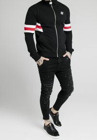 Chaqueta negra con cremallera y franjas rojas y blancas en las mangas; combinada con jeans negros desgastados que tienen manchas de pintura; zapatillas negras.