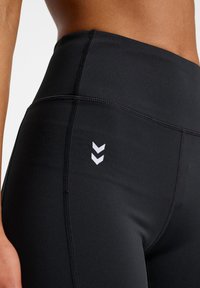 Czarne legginsy treningowe wykonane z elastycznego materiału, z wysokim stanem i białym logo w kształcie zygzaka na dolnej części. Widoczne detale szwów.