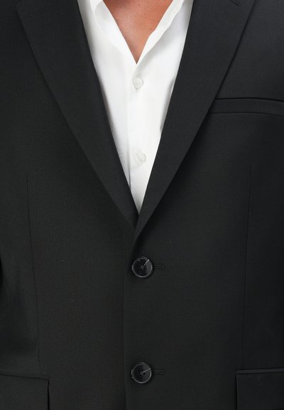Veste noire avec revers crantés, poche poitrine simple et trois boutons. Portée sur une chemise blanche à col. Tissu lisse.
