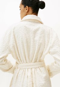 Vestaglia color crema con motivi floreali e paisley testurizzati, caratterizzata da un colletto a scialle e una cintura da annodare, realizzata in un tessuto morbido e soffice.