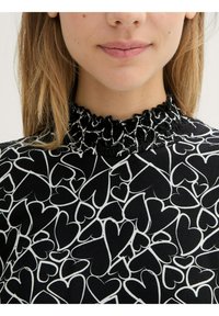 Zwarte blouse met een wit hartpatroon, voorzien van een hoge, gerimpelde halslijn en een gestructureerde stof. Close-up van het halsgedeelte.