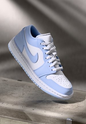 Baskets basses Nike blanches et bleu clair, avec des lacets, des perforations sur la boîte à orteils et le logo Swoosh emblématique, présentées au-dessus d'une surface en béton.