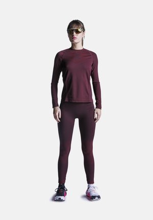 Vrouw staand, gekleed in een bordeauxrode top met lange mouwen en legging, reflecterende sportzonnebril en niet-overeenkomende zwarte en witte hardloopschoenen.