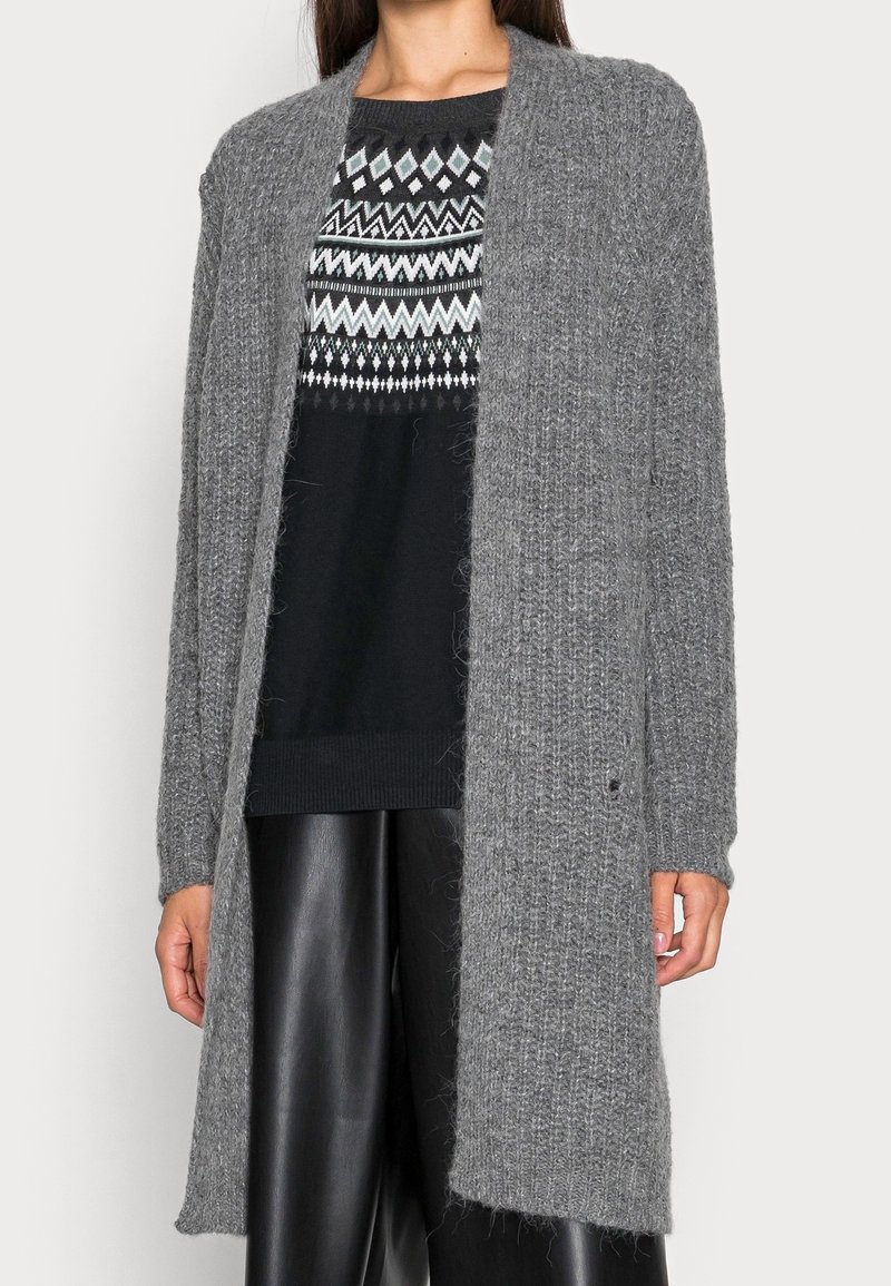 Cardigan long gris en maille texturée, avec un devant ouvert et des poignets côtelés, porté sur un pull noir à motif.
