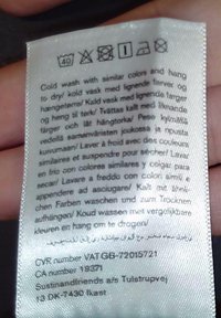Ετικέτα φροντίδας ρούχων με πολύγλωσσες οδηγίες για πλύσιμο σε κρύο νερό και στέγνωμα με κρέμασμα, κρατημένη μεταξύ των δαχτύλων.