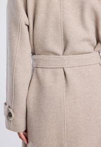 Abrigo beige de espiga con cintura ceñida y puños abotonados. Presenta un diseño estructurado y una textura suave.