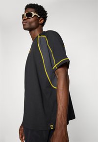 Camisa preta de manga curta com detalhes em costura amarela e um tecido texturizado. Apresenta uma etiqueta do logotipo na bainha e um design de corte relaxado.