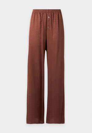 Femme aux longs cheveux blonds porte un haut beige sans manches, un pantalon ample couleur rouille, des chaussures noires, et tient un sac à main vert foncé dans une main.