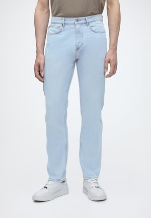 Homme portant un jean droit bleu clair et des baskets blanches, debout devant un fond uni.