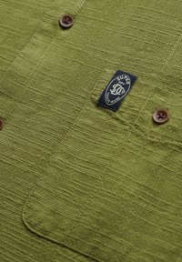 Camisa verde texturizada con un patrón de tejido, que presenta una etiqueta azul y botones marrones. La tela parece suave y transpirable.