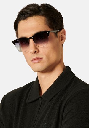 Gafas de sol con montura negra y lentes degradados. Forma redonda, varillas finas y detalles metálicos. El modelo lleva una chaqueta negra con cremallera y textura.