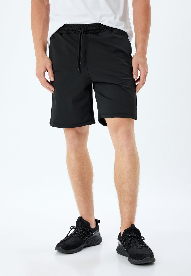 Pantalones cortos deportivos negros con cintura elástica y cordón, textura suave, de longitud por encima de la rodilla, combinados con zapatillas deportivas negras.