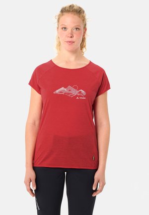 Junge Frau mit lockigen blonden Haaren, die ein rotes Vaude-T-Shirt mit weißem Bergmotiv und schwarze Hose trägt, stehend vor einem weißen Hintergrund.