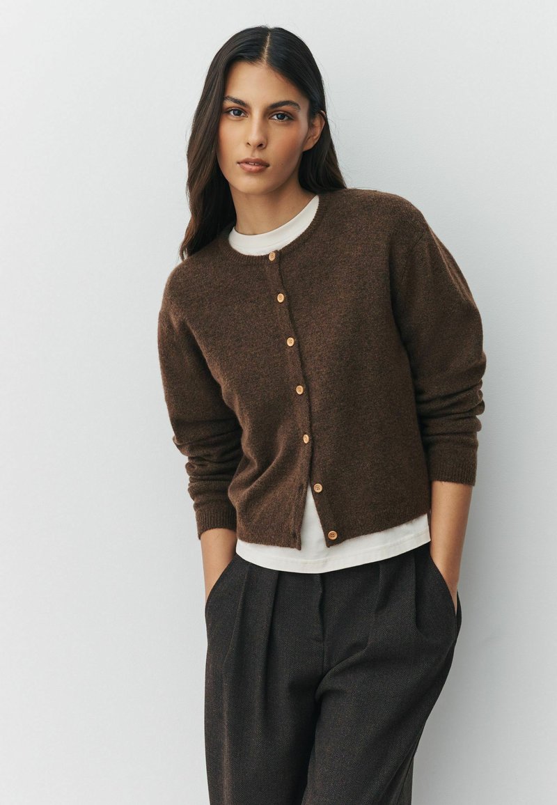 Hnědý pletený kardigan s kulatým výstřihem a dřevěnými knoflíky. Nosený přes bílou košili, v kombinaci s tmavými skládacími kalhotami.